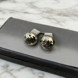 Brincos de metal dourado com esfera texturizada e detalhes escuros, sobre base cinza