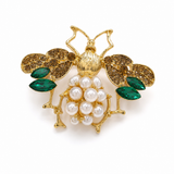 Broche dourado em forma de abelha rainha, com pedras verdes e pérolas brancas.