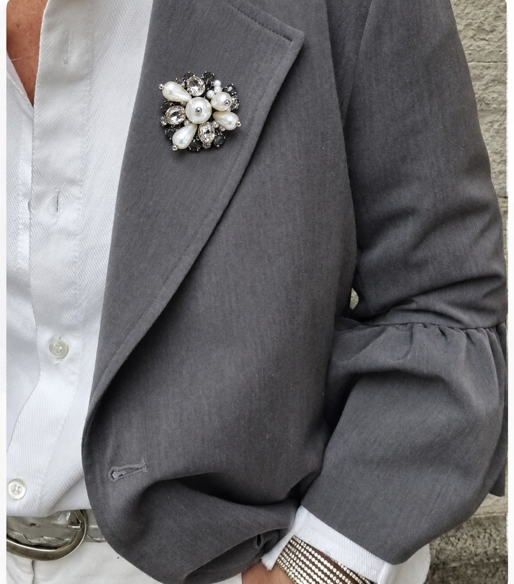 Broche de pérolas e strass com pedras escuras, preso na lapela de blazer cinza