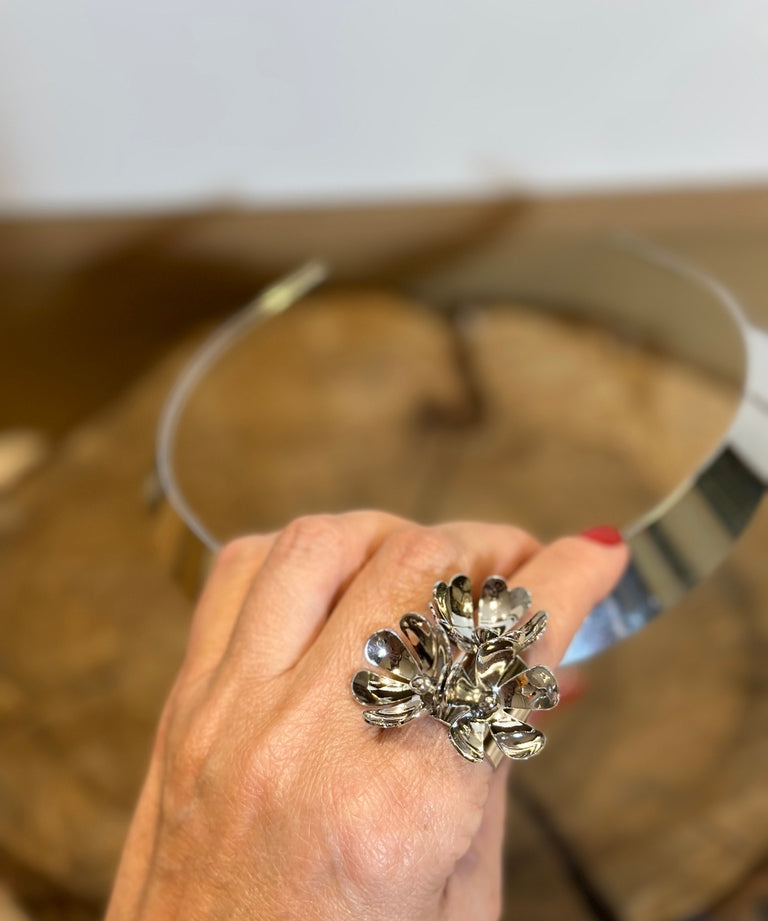 Anel prateado em formato de flor, com miolo escuro e pétalas brilhantes.