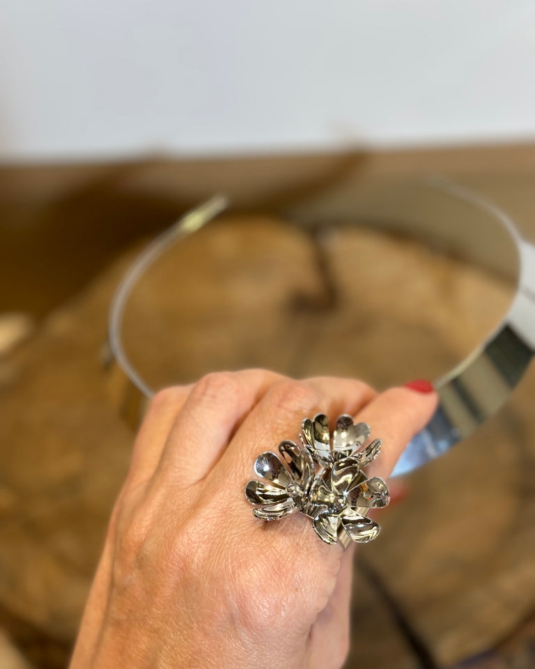 Anel prateado em formato de flor, com miolo escuro e pétalas brilhantes.