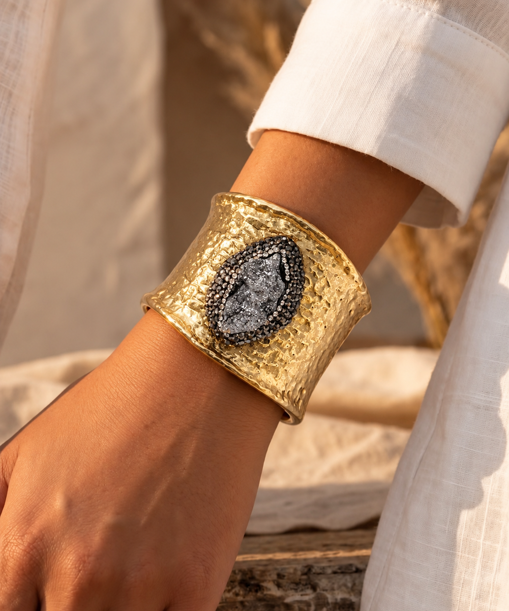 Bracelete Fira largo dourado com pedra oval cinza-escura, usado no pulso