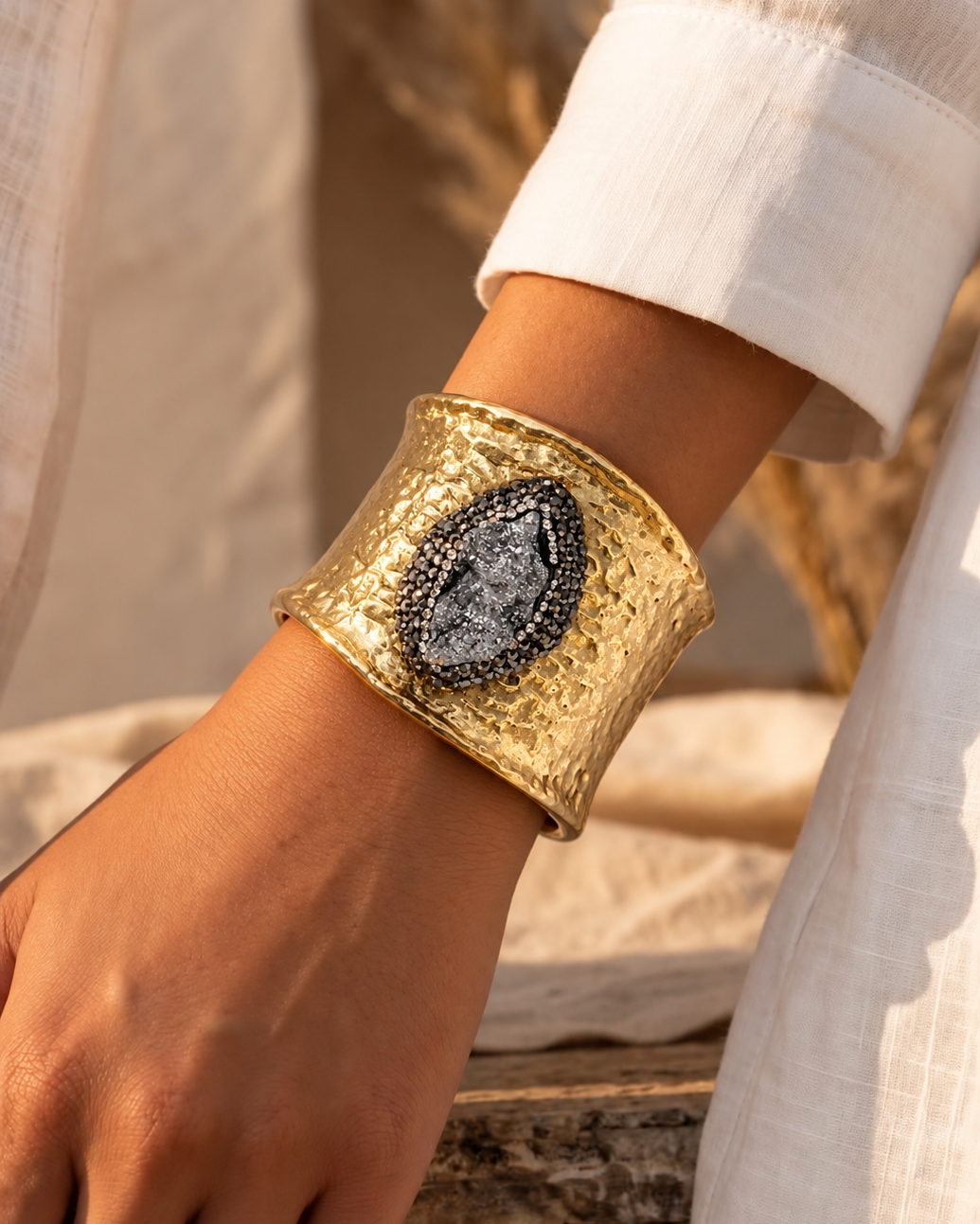 Bracelete Fira largo dourado com pedra oval cinza-escura, usado no pulso