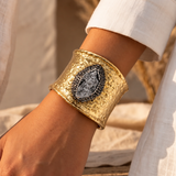 Bracelete Fira largo dourado com pedra oval cinza-escura, usado no pulso