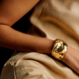 Bracelete dourado espelhado em mão, textura martelada, sobre tecido bege