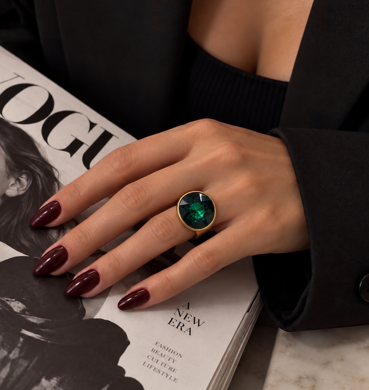 Anel dourado com pedra redonda verde na mão, unhas vinho
