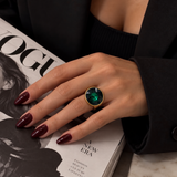 Anel dourado com pedra redonda verde na mão, unhas vinho