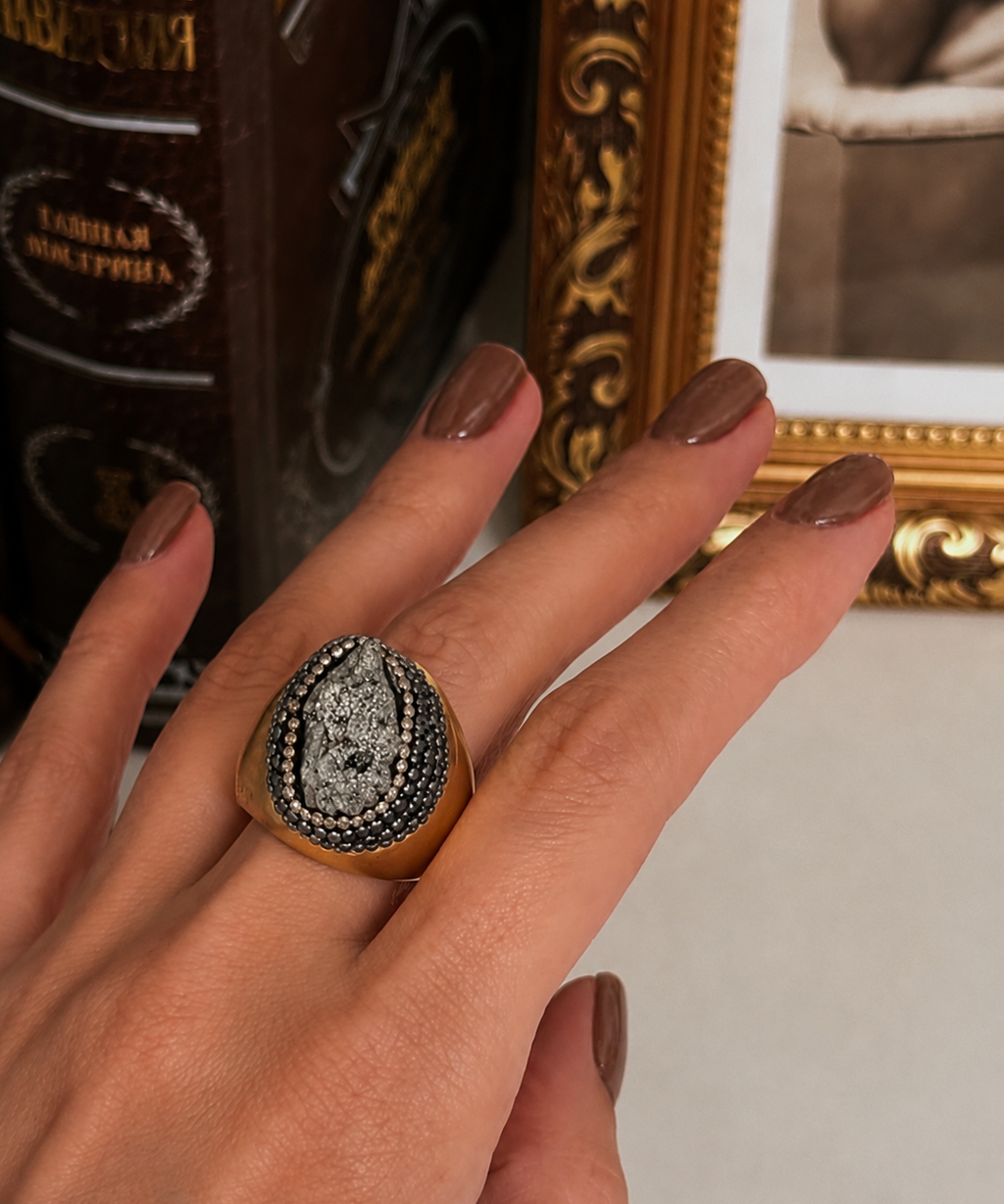 Anel Fira dourado com pedra oval cinza-escura e moldura cravejada