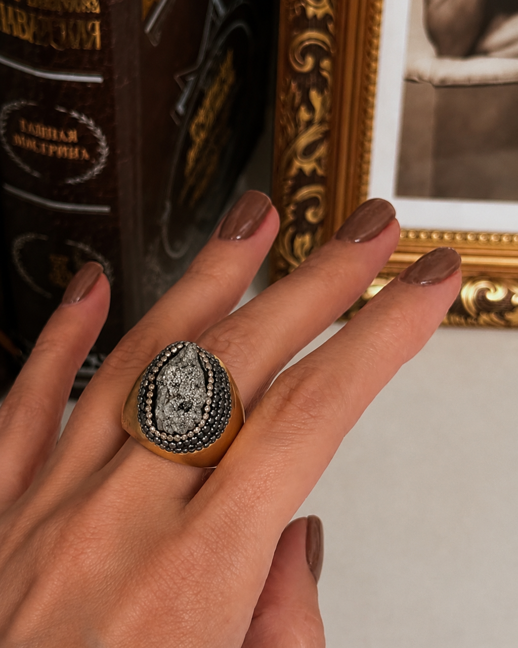 Anel Fira dourado com pedra oval cinza-escura e moldura cravejada
