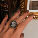 Anel Fira dourado com pedra oval cinza-escura e moldura cravejada