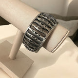 Bracelete metálico largo com padrão gravado em linhas e recortes, acabamento escuro