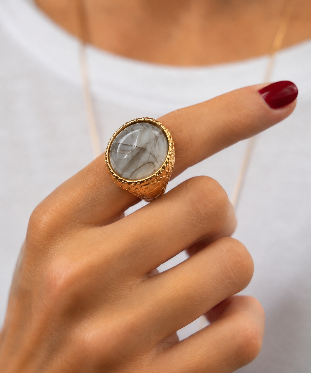 Anel dourado com pedra oval transparente com veios, usado em mão